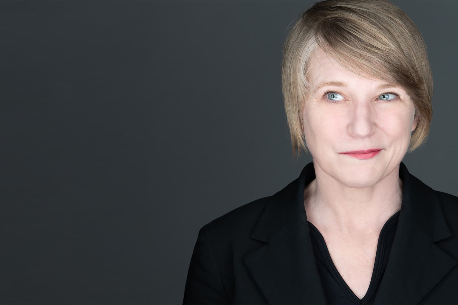 An actor headshot of a women wearing a black top, with short hair, on a gray background