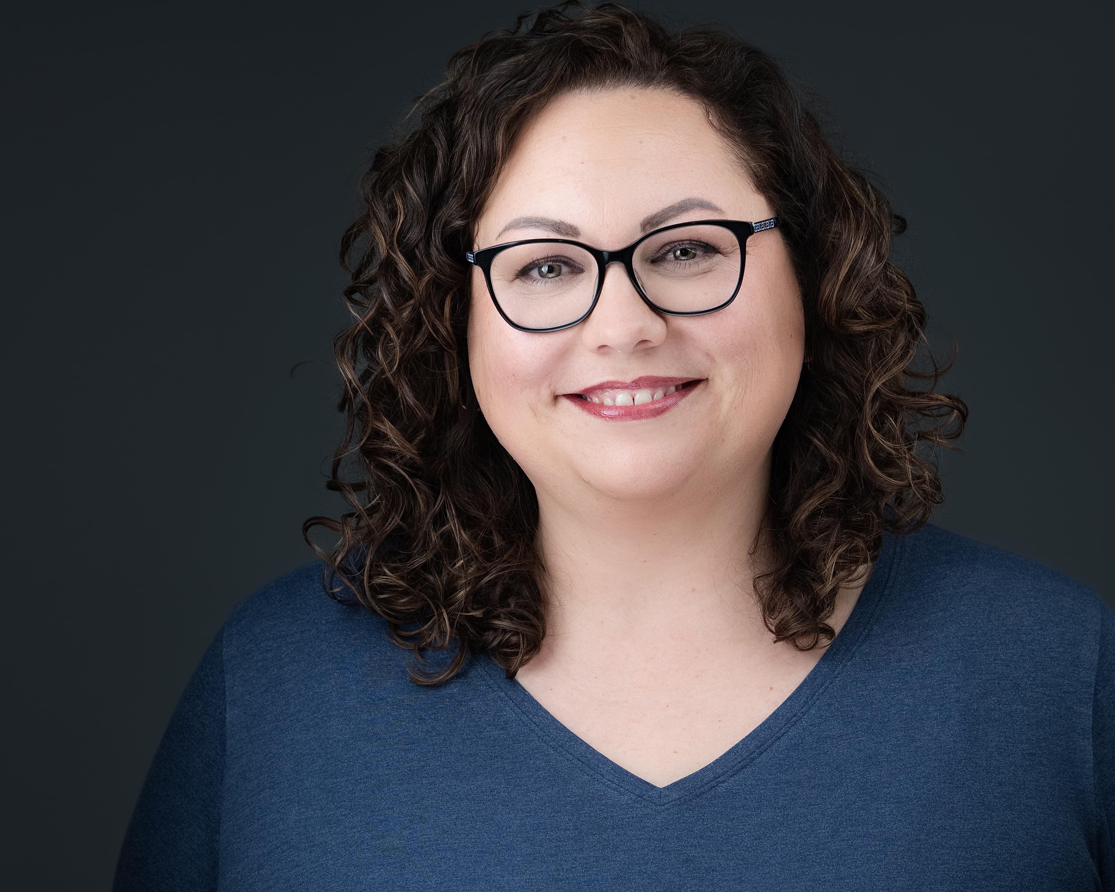 Woman with glasses and curly hair wearing a blue shirt against a dark background