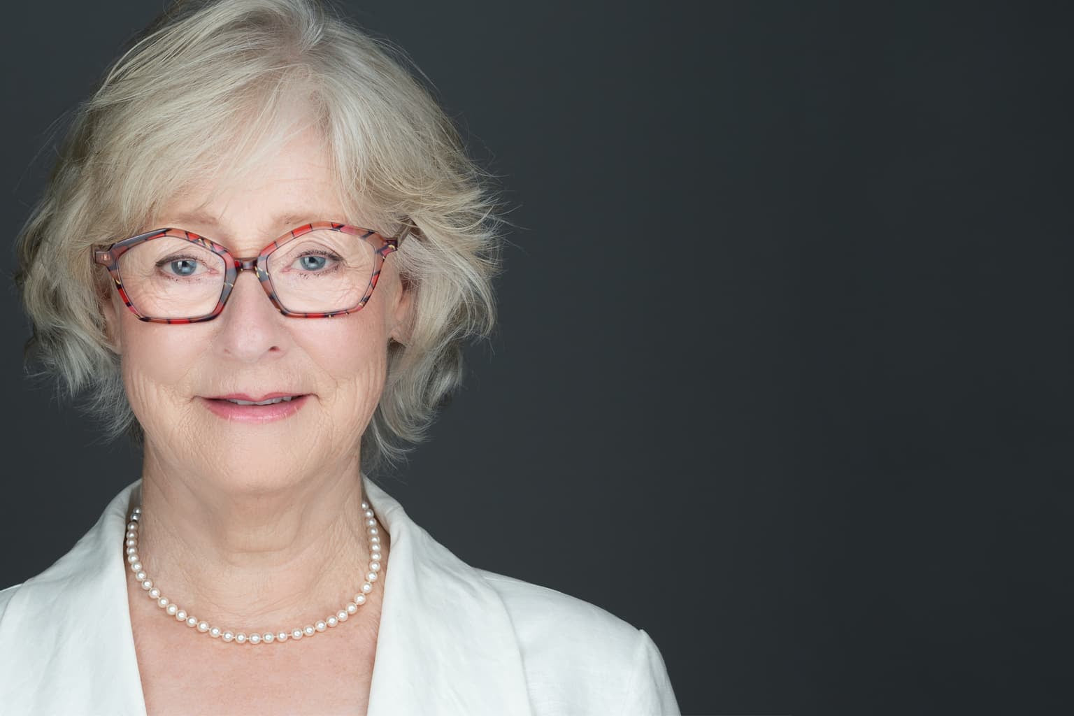 Author headshot of a women wearing a white jacket, red glasses, pearl necklace, on a gray background