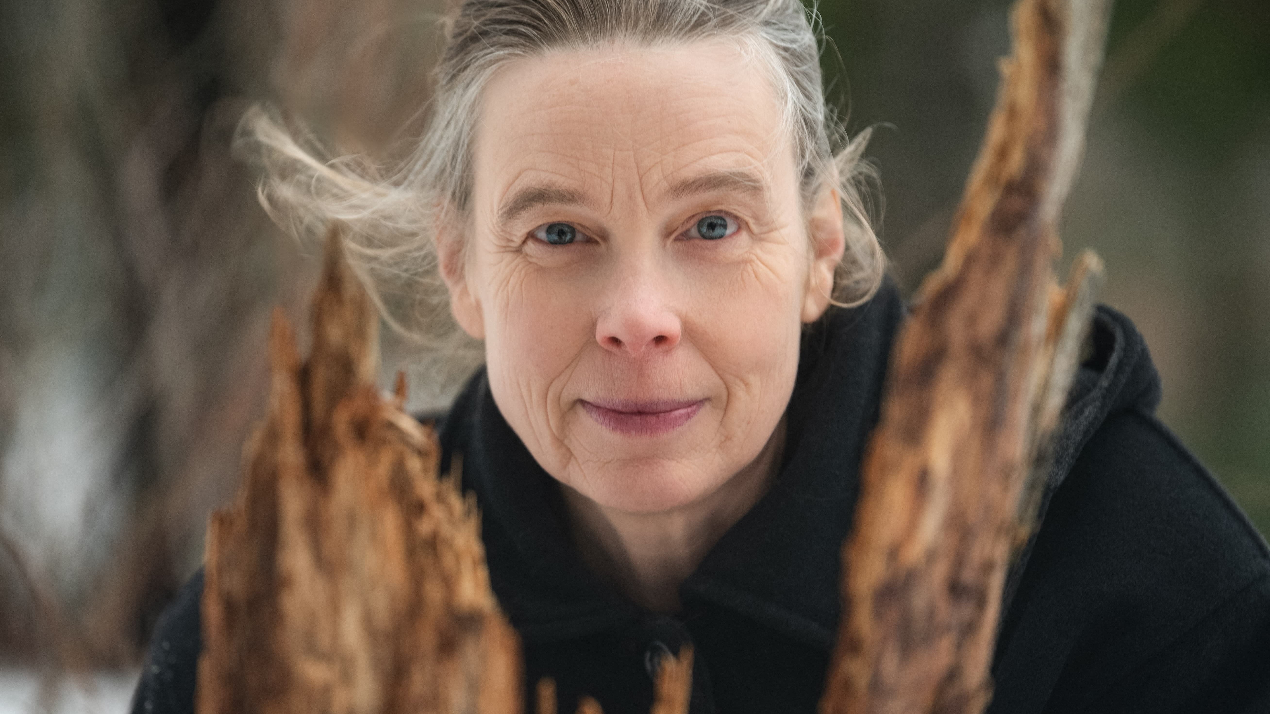 Person with gray hair standing between two large wooden logs in a natural setting