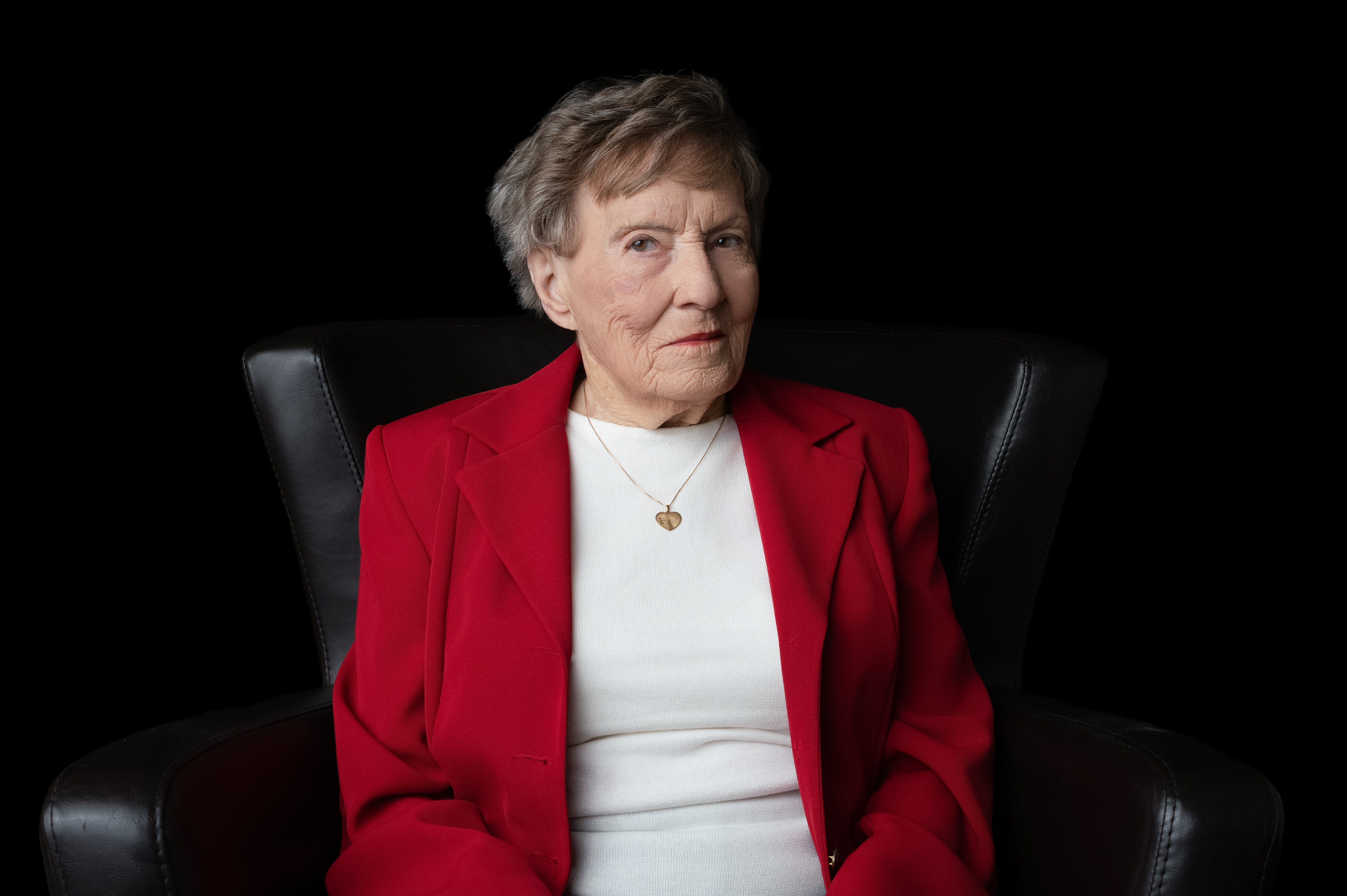 Ellen Gilchrist portrait in bright red blazer and white top against black studio backdrop, seated with dignified posture