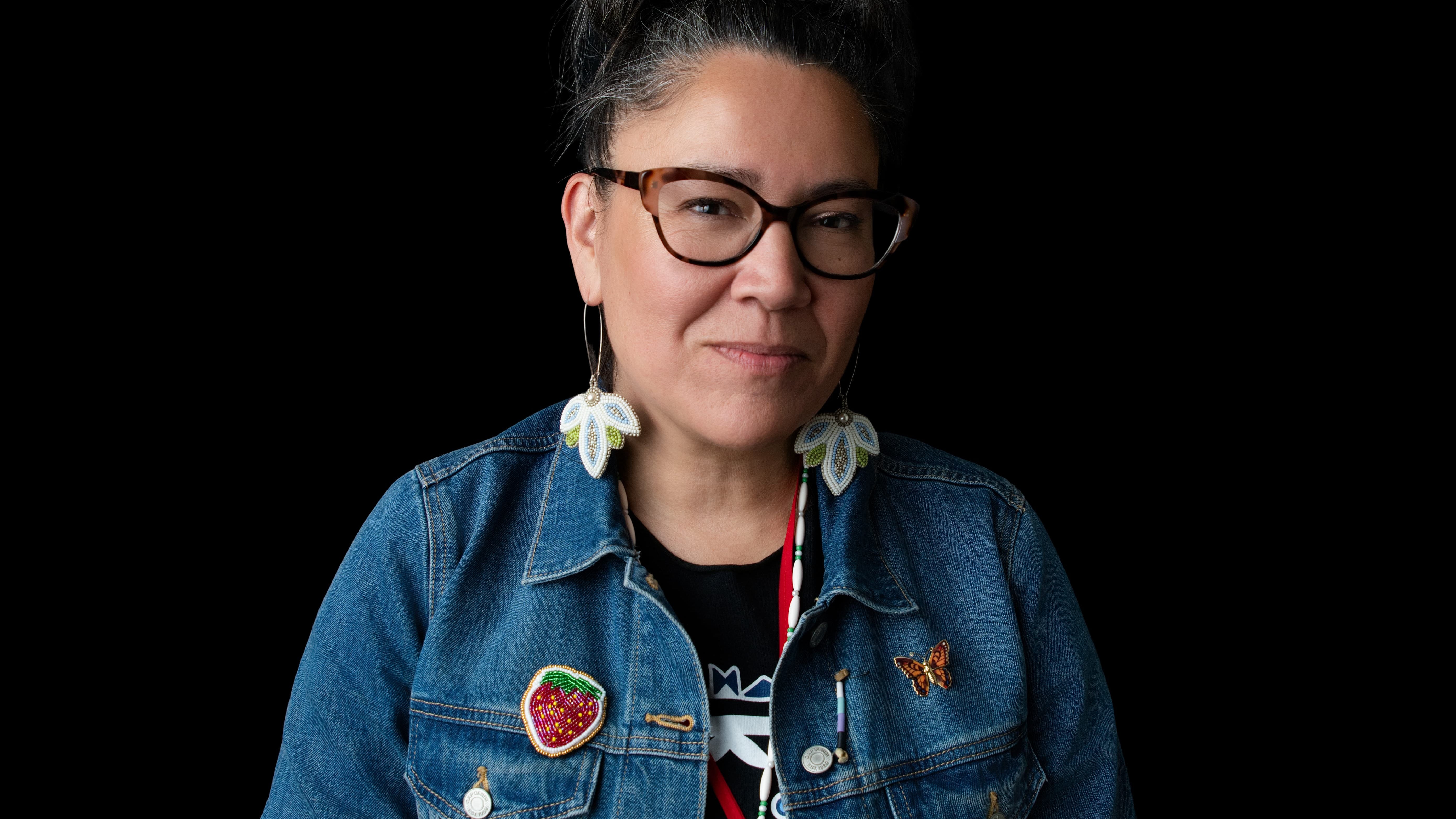 A portrait of a woman with glasses and dark hair pulled up, wearing a blue denim jacket adorned with various colorful patches, including a strawberry patch and a butterfly patch, against a solid black background.