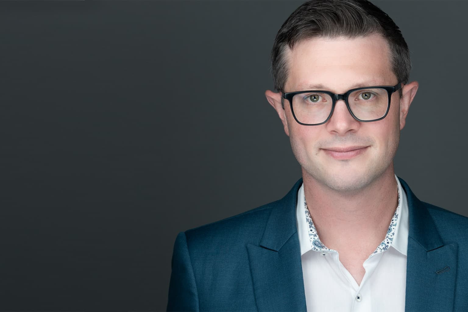 Business headshots of a young man who has glasses, wearing a white shirt and blue sports jacket