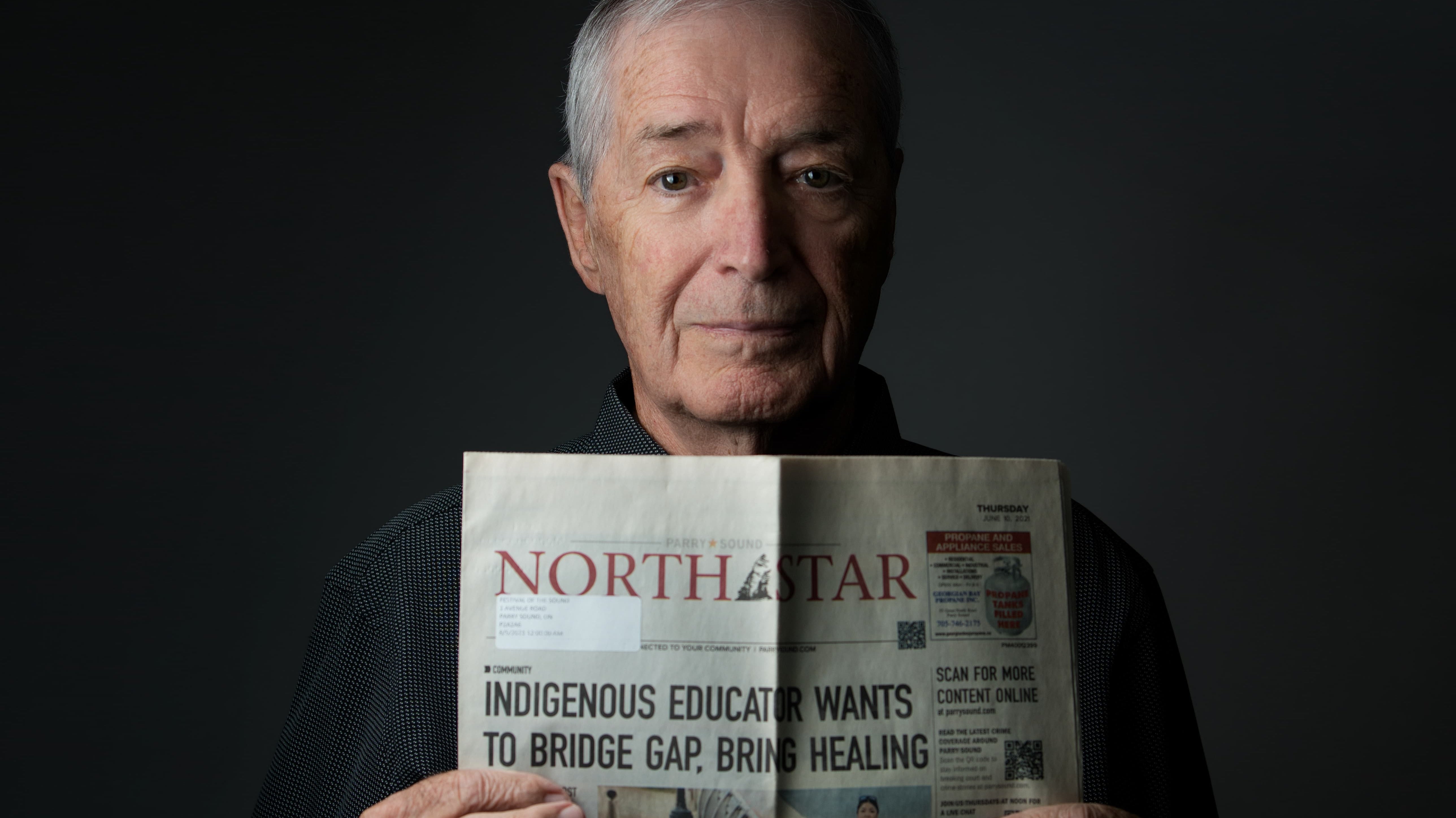 An older man holding a folded North Star magazine against a dark studio background. The visible front cover headline reads, 'INDIGENOUS EDUCATOR WANTS TO BRIDGE GAP, BRING HEALING.