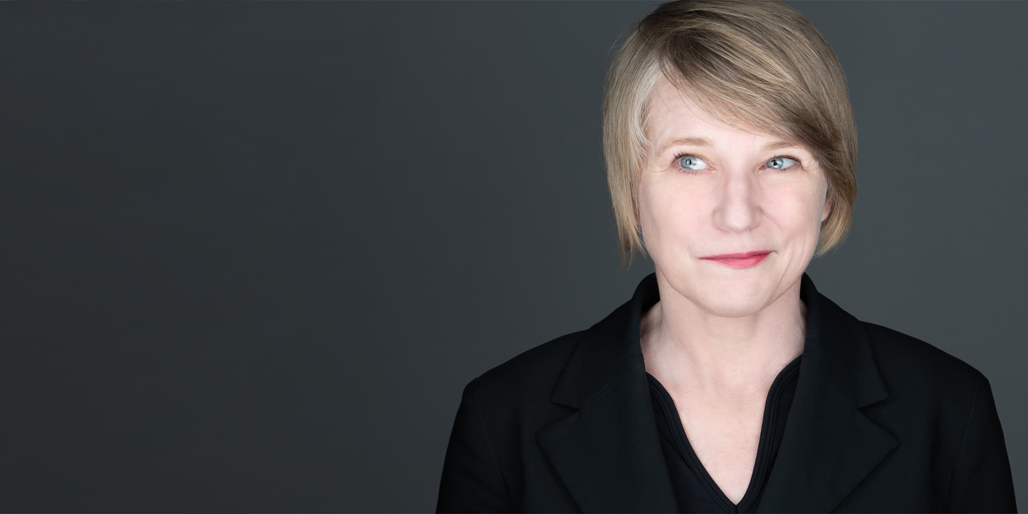An actor headshot of a women wearing a black top, with short hair, on a gray background