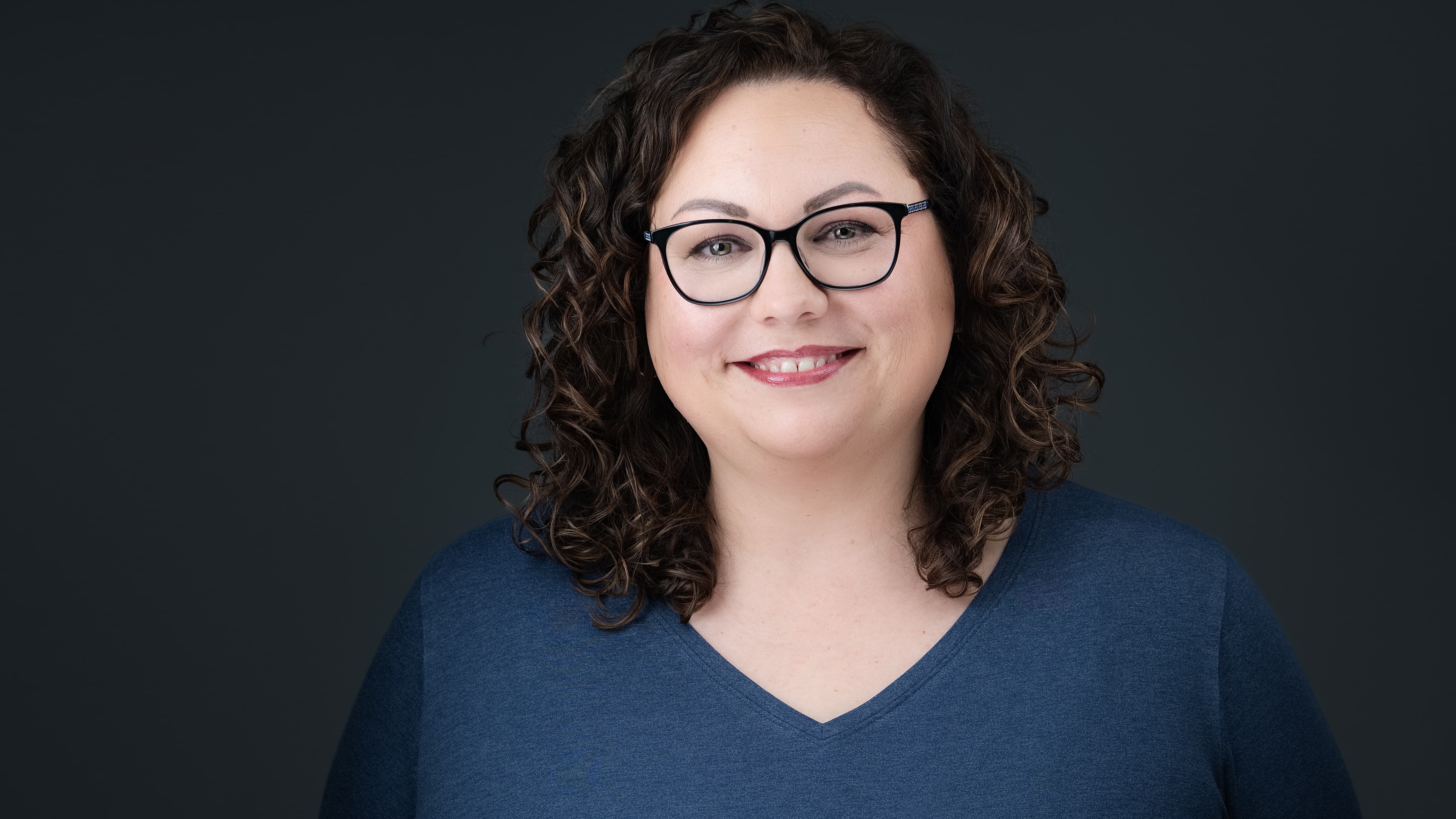 Woman with glasses and curly hair wearing a blue shirt against a dark background