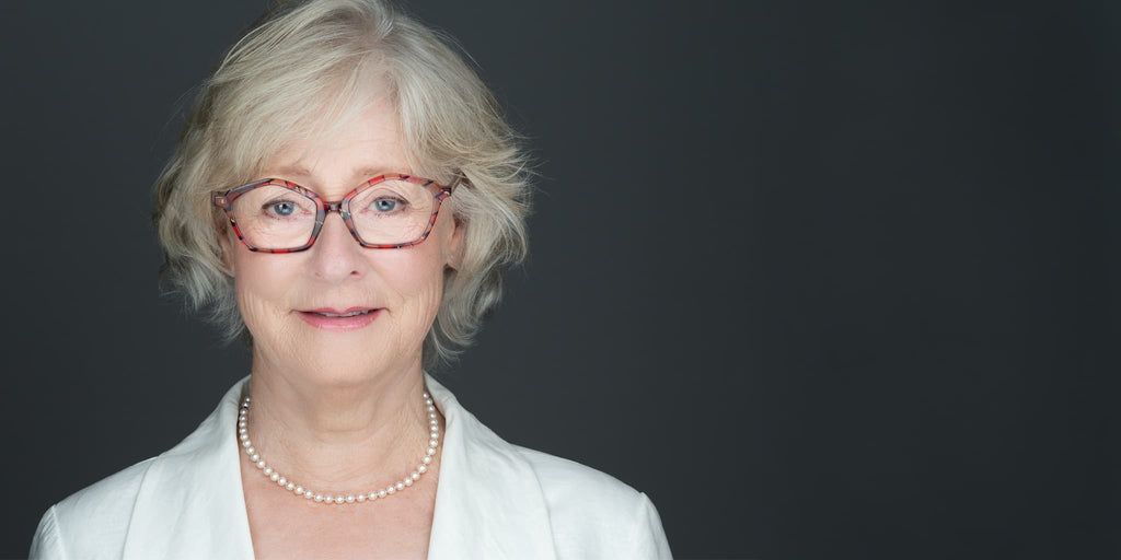 Author headshot of a women wearing a white jacket, red glasses, pearl necklace, on a gray background