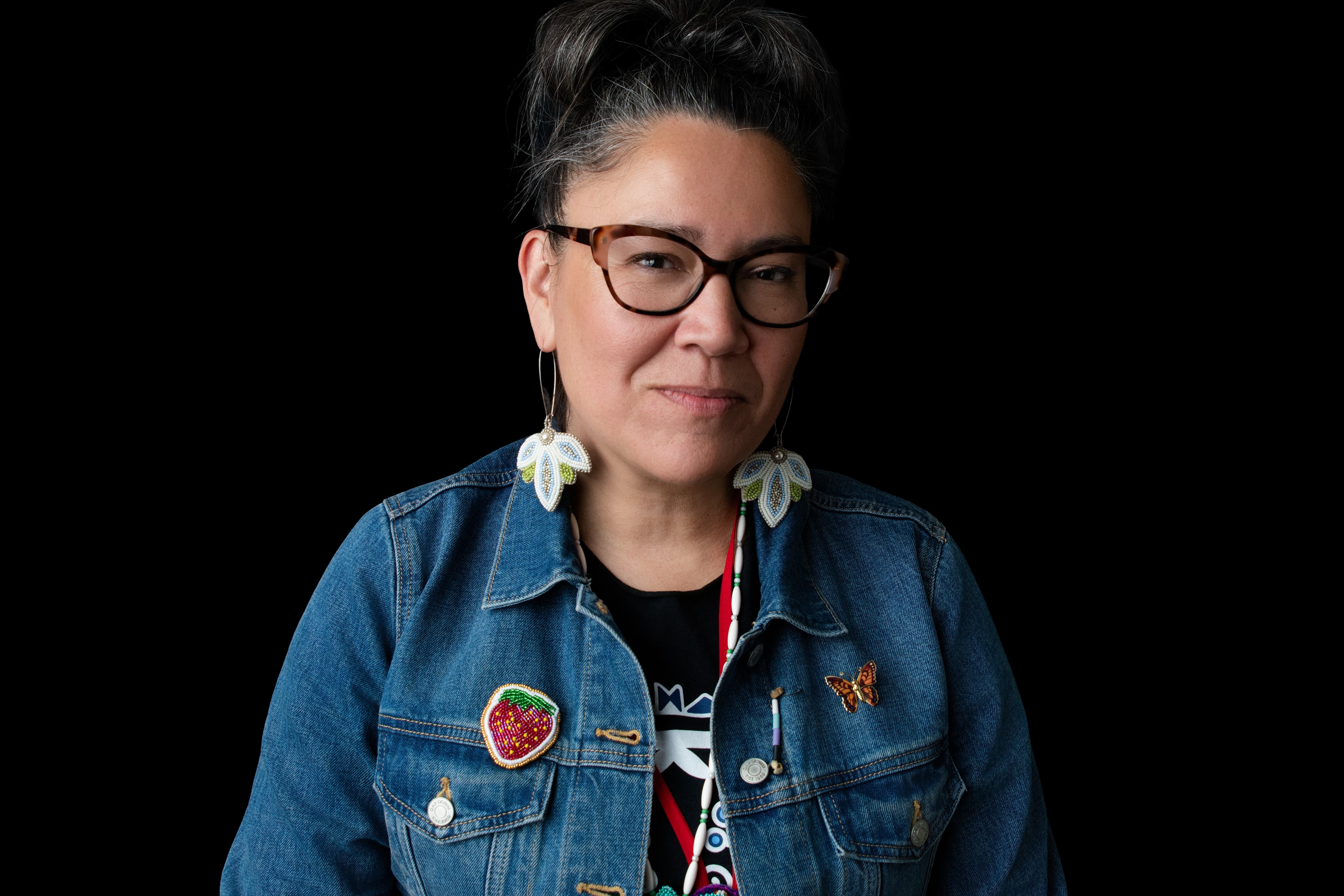A portrait of a woman with glasses and dark hair pulled up, wearing a blue denim jacket adorned with various colorful patches, including a strawberry patch and a butterfly patch, against a solid black background.