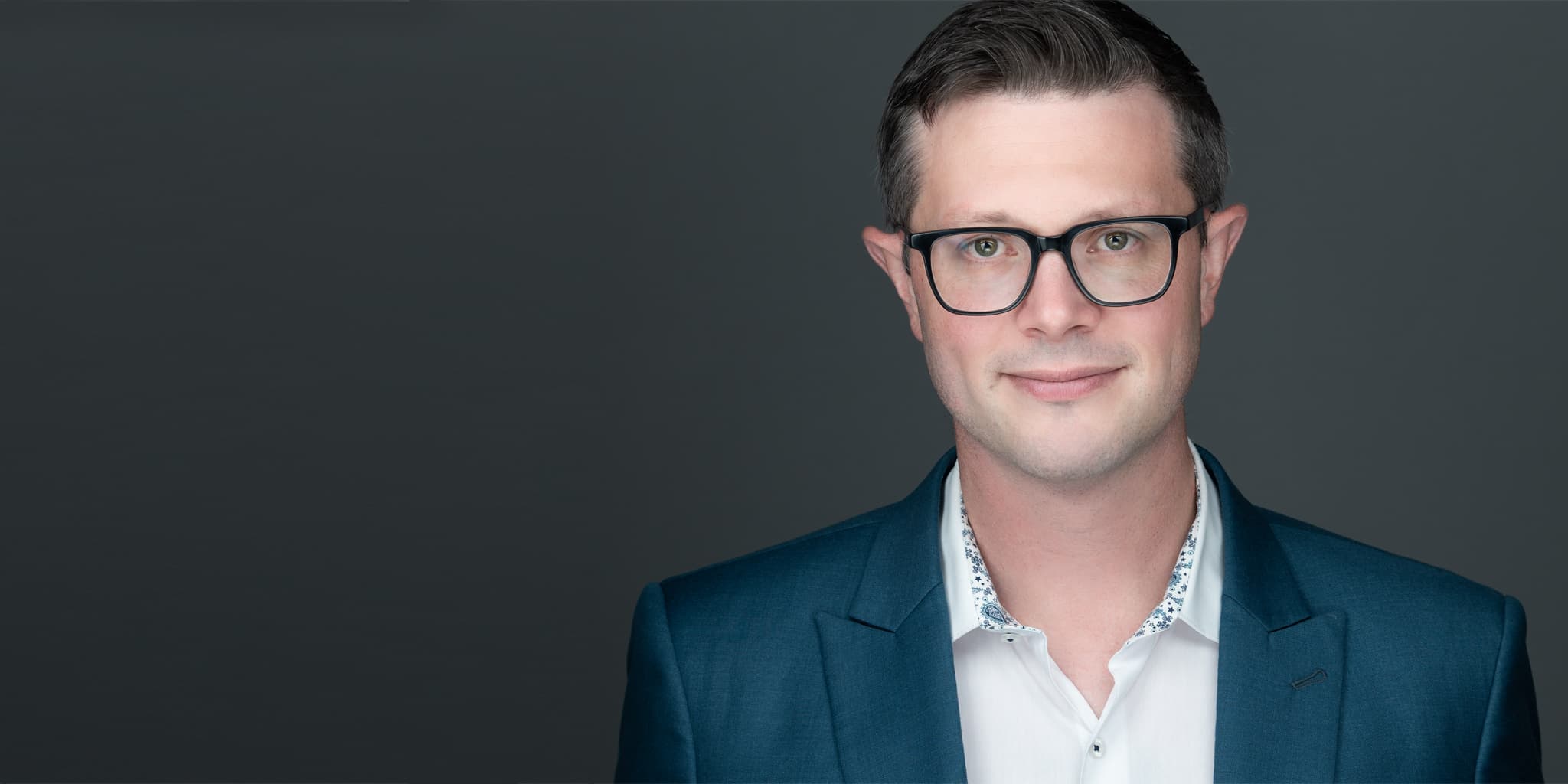 Business headshots of a young man who has glasses, wearing a white shirt and blue sports jacket
