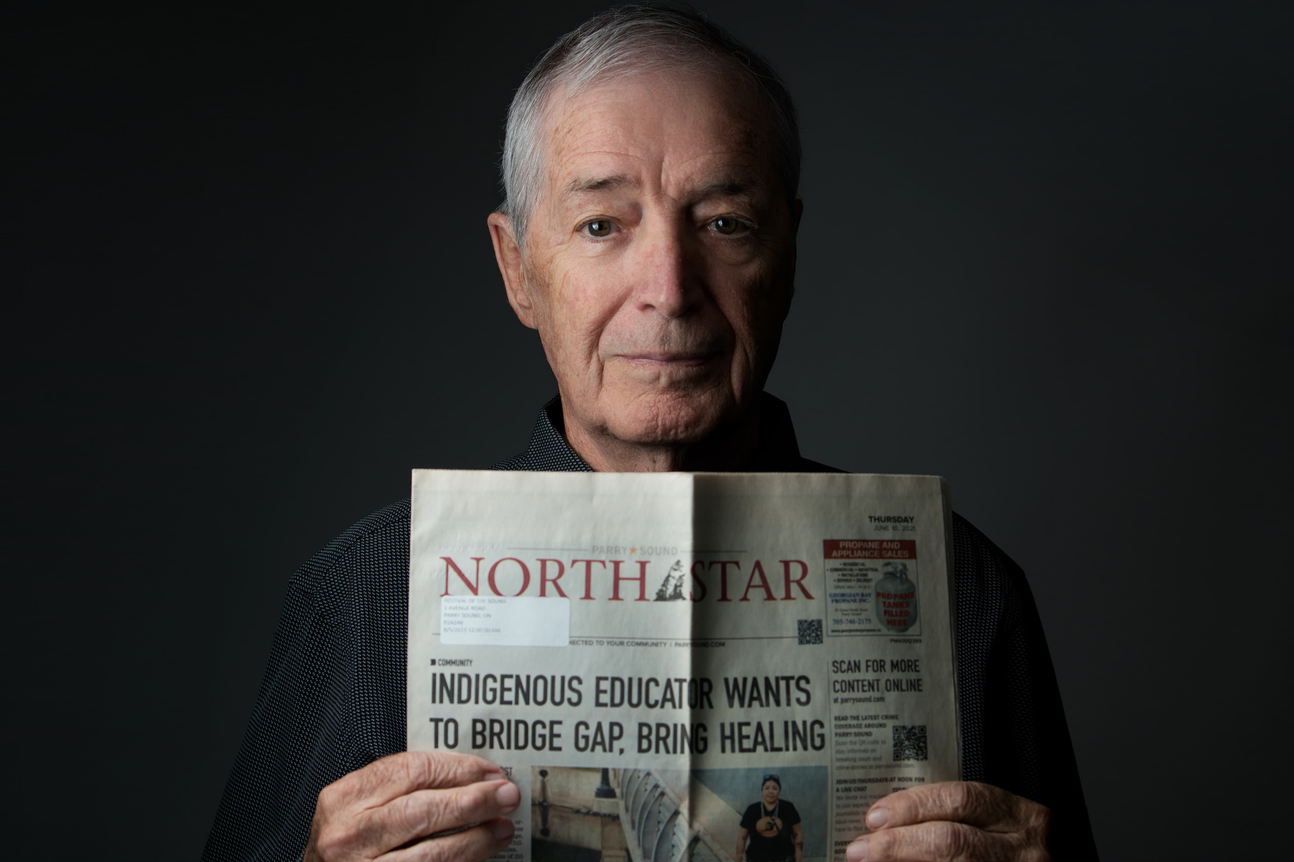 An older man holding a folded North Star magazine against a dark studio background. The visible front cover headline reads, 'INDIGENOUS EDUCATOR WANTS TO BRIDGE GAP, BRING HEALING.